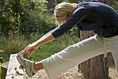 Young woman in a forest stretching her leg over a wooden beam and touching her foot\n