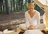 Female massage therapist sitting behind a woman massaging her scalp under a tent in a forest\n