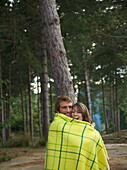 Young couple standing in a forest wrapped in a blanket laughing\n