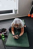 Senior woman using phone while stretching on mat in gym\n