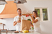 Smiling couple with paper shopping bag and groceries in kitchen\n