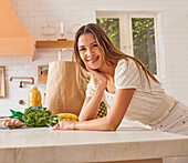 Portrait of smiling woman with paper shopping bag and groceries in kitchen\n