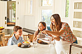 Smiling mother with son (8-9) and daughter (12-13) having breakfast in kitchen\n
