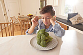 Unzufriedener Junge (8-9) betrachtet den Brokkoli auf dem Teller in der Küche