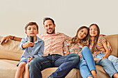 Smiling family with two children (8-9, 12-13) watching TV on sofa\n