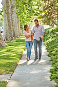 Lächelndes Paar hält sich an den Händen, auf einem mit Bäumen gesäumten Bürgersteig gehend
