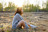 Side view of woman listening to music while sitting on grass in meadow at sunset\n