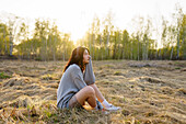 Seitenansicht einer Frau, die Musik hört, während sie im Gras auf einer Wiese bei Sonnenuntergang sitzt