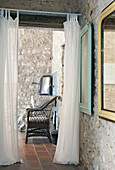 View through open curtains to rattan armchair in room with natural stone walls