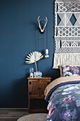 Macramé wall hanging over vintage bedside table in bedroom with dark blue wall