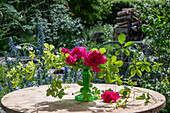 Rote Rosenblüten (Rosa) dekoriert mit Stein auf Stielvase und Zweigen auf Gartentisch