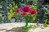 Rote Rosenblüten (Rosa) dekoriert mit Stein auf Stielvase und Zweigen auf Gartentisch