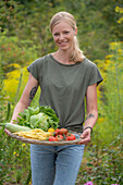 Junge Frau trägt Tablett aus Korb mit frisch geerntetem Gemüse, Eisbergsalat, Zucchini, Tomaten und Bohnen 'Neckargold' (Phaseolus)