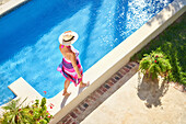 Woman in straw hat walking along edge of sunny swimming pool