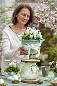 Frau baut Etagere mit Blumentopf aus Hornveilchen, Gänseblümchen und Glasvase, Ostereier in kleinem Moosnestern mit Federn und auf Tisch