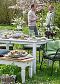 Junges Paar vor gedecktem Tisch, Osterfrühstück mit Osternest und gefärbten Eiern in Eierbechern, Narzissen und Petersilie im Korb im Garten