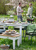 Young couple behind table laid for Easter breakfast with Easter nest and colored eggs in egg cups, daffodils and parsley in a basket in the garden