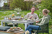 Young couple sitting at a laid table for Easter breakfast with Easter nest, colored eggs, cereal bowls, bouquet of flowers and water jug in the garden