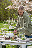 Junge Frau baut Etagere aus Blumenstrauß auf Suppenschüssel, gedeckter Tisch zum Osterfrühstück mit Osternest und gefärbten Eiern und Wasserkrug
