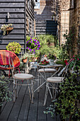 Kleiner Sitzbereich mit Metallstühlen und -tisch auf einer Holzterrasse, umgeben von blühenden Pflanzen