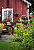 Idyllische Gartenecke vor roter Hauswand mit blühenden Pflanzen und Lichterkette