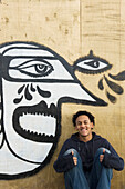 UK,Teenage boy sitting in front of graffiti wall of cubist style face,Hastings