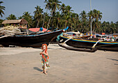 Isla Reynolds, 4 Jahre alt, steht vor den vielen traditionellen Booten und Strandrestaurants, die den Strand von Palolem säumen, Goa, Indien.