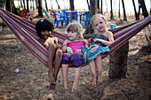 Britische Ferienkinder spielen in einer Hängematte und freunden sich mit einem einheimischen Mädchen am Turtle Beach, Goa, Indien, an.