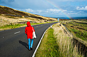 United Kingdom,England,Woman in red coat walking along A635 Greenfiled road. Rainbow in distance,Yorkshire