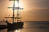 Silhouette von Menschen an Bord eines großen Schiffes, das am Dock in der Hauptstadt der Kaimaninseln, George Town, vertäut ist, mit Kajakfahrern an der Seite, die im goldenen Schein der Dämmerung aufs Meer hinauspaddeln, George Town, Grand Cayman, Kaimaninseln, Karibik