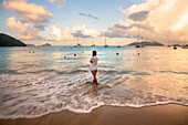 Blick von hinten auf eine Frau, die in der schäumenden Brandung am Strand steht und auf das türkisfarbene Wasser mit Booten schaut, die vor dem Ufer entlang des Horizonts in der Cane Garden Bay in der Abenddämmerung vertäut sind, Tortola, Britische Jungferninseln, Karibik