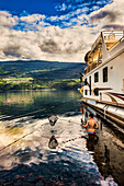 Eine Familie genießt ihren Urlaub auf einem Hausboot, das am Ufer des Shuswap Lake geparkt ist, während ein Mann vom Deck des Hausboots aus angelt und ein Junge im Badeanzug im Wasser steht und ein Fischernetz bereithält, Shuswap Lake, British Columbia, Kanada
