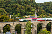 Elektrischer Zug auf alter Steinbogenbrücke mit Kirchturm und bewaldeten Hügeln im Hintergrund, Luxemburg-Stadt, Luxemburg