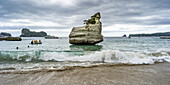 Die berühmte Cathedral Cove, die nur zu Fuß oder mit dem Boot zu erreichen ist, gehört zu den wichtigsten Sehenswürdigkeiten auf dem Coromandel. Schwimmer genießen es, in der Brandung zu spielen, während Boote mit Touristen ankommen, Waikato Region, Neuseeland