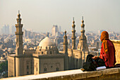 Eine junge Frau sitzt mit einer Kamera auf einer Mauer in der Großen Moschee von Muhammad Ali Pascha und blickt auf die Moschee-Madrassa von Sultan Hassan und das Stadtbild von Kairo, Kairo, Ägypten