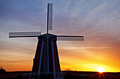 Windmühle bei Sonnenaufgang mit einer goldenen Sonne, die über dem Horizont aufgeht, und dem Gipfel des Mount Hood als Silhouette in der Ferne, Willamette Valley, Oregon, Vereinigte Staaten von Amerika