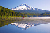 Spiegelbild des Mount Hood und des Waldes, der sich im Trillium Lake spiegelt, mit vom Wasser aufsteigendem Nebel und Menschen, die in dem ruhigen Wasser Boot fahren und angeln, Oregon, Vereinigte Staaten von Amerika