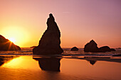 Rugged rock formations along the shoreline with a bright sun glowing in the sky at sunrise and reflecting on the wet sand at Bandon State Natural Area on the Oregon coast,Bandon,Oregon,United States of America