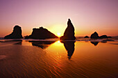 Zerklüftete Felsformationen entlang der Küste mit einer hellen Sonne, die bei Sonnenaufgang am Himmel leuchtet und sich auf dem nassen Sand in der Bandon State Natural Area an der Küste von Oregon, Bandon, Oregon, Vereinigte Staaten von Amerika spiegelt