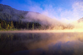 Niedrige Wolken und Nebel über dem Trillium Lake bei Sonnenaufgang mit einem dichten Wald, der sich im Wasser spiegelt, Mount Hood National Forest, Oregon, Vereinigte Staaten von Amerika