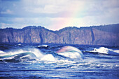 Plätschernde Wellen mit einem Regenbogen, der aus dem Wasser in den Himmel ragt, entlang der Küste von Cape Kiwanda an der Küste von Oregon in Cape Kiwanda State Natural Area, Oregon, Vereinigte Staaten von Amerika