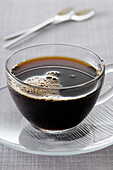 Nahaufnahme von schwarzem Kaffee in Glastasse und Untertasse vor grauem Hintergrund, Studioaufnahme