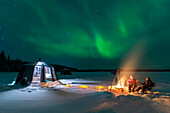 Nachtansicht von zwei Personen, die draußen am Lagerfeuer in der Nähe des beleuchteten Glas-Iglus mit tanzenden Nordlichtern (Aurora Borealis) am Himmel zu Abend essen, Jokkmokk, Norrbotten, Schwedisch-Lappland, Schweden, Skandinavien, Europa