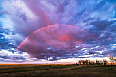 A nearly semi-circular rainbow that appeared briefly right at sunset so the warm lighting made the rainbow appear more red than usual and set amid red clouds,brighter within the rainbow than outside the bow.