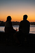 Ein Paar genießt einen Drink bei Sonnenuntergang auf der Promenade Jacques Thibaud vor dem Strand Grande Plage in Saint Jean de Luz, einem Fischerdorf an der Mündung des Flusses Nivelle im südwestlichen Baskenland in Frankreich