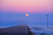 Die untergehende Sonne, die auf einer Landstraße in eine Nebelbank eintaucht, mit Blickrichtung Westen, am Abend des Frühlingsäquinoktiums, 20. März 2023. Die Sonne geht also in Richtung Westen unter. Der Nebel verdunkelt und rötet die Sonne, was die atmosphärische Absorption veranschaulicht. Dies war auf dem Highway 561 in Süd-Alberta.