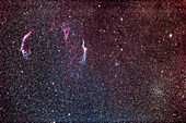 The arcs and wisps of the Veil Nebula supernova remnant in Cygnus,framed with the open star cluster NGC 6940 in Vulpecula at lower right. The area is marked by a sharp transition between clear starry sky in the Millky Way and brownish-yellow areas obscured by interstellar dust with fewer stars visible. The nebula colours show up well despite this being a "stock" camera,with no filters employed.