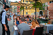 Ein Mann fährt mit einer Gondel an einer Restaurantterrasse an einem Kanal in Venedig, Italien, vorbei. Touristen speisen in einem beliebten Ristorante im Freien in der Fondamenta de l'Osmarin, San Marco, Venedig, Italien, entlang eines Kanals mit Gondeln und Gondoliere
