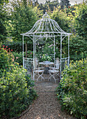 Weiße Ranklaube mit Metalltisch und Stühlen im üppigen Garten