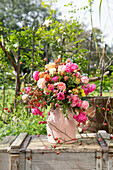 Bouquet of roses with rose hips in pink ceramic vase on wooden box in the garden